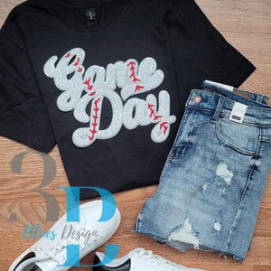 May include: Black t-shirt with "Game Day" text in a baseball design, paired with denim shorts and white sneakers. The text is in a baseball stitch design with red accents. A casual outfit for game day.