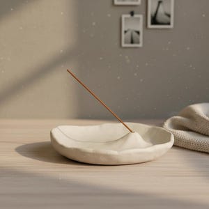 May include: A cream-colored ceramic incense holder with a raised center, holding a brown incense stick. The holder sits on a light wood surface, with a folded beige blanket in the background. Decorative wall art is visible.