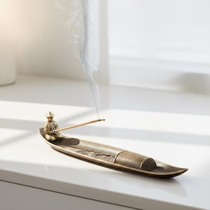 Peut inclure: Un porte-encens en bronze avec une figurine assise et un design en forme de bateau. Le support présente un motif texturé et dégage de la fumée d'un bâton d'encens allumé. Le fond est une surface blanche et une fenêtre.