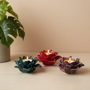 May include: Three ceramic lotus flower candle holders in teal, red, and purple, each featuring a glossy finish and layered petals. A small lit tea light candle sits in the centre of each holder, casting a warm glow from the flame.