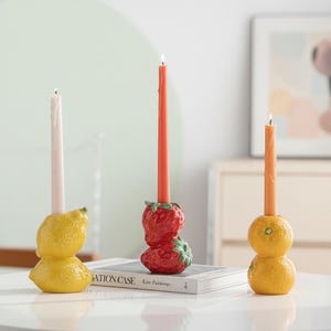 Puede incluir: Tres candelabros decorativos con velas encendidas. Uno es amarillo y tiene forma de limón, otro es rojo y tiene forma de fresa, y el tercero es amarillo y tiene forma de naranja. Las velas son rosas, rojas y naranjas.