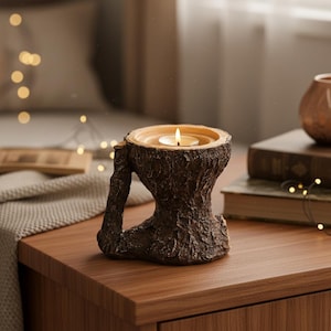 Puede incluir: Un portavelas marrón con forma de tronco de árbol con una vela de té encendida. El portavelas tiene una superficie texturizada y una extensión en forma de asa. El fondo incluye una superficie de madera, libros y una iluminación suave.