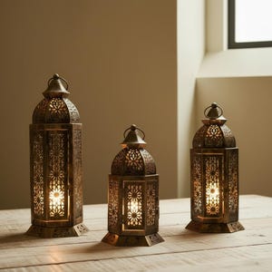 Op de afbeelding: Drie bronzen metalen lantaarns met ingewikkelde bloemmotieven. Elke lantaarn heeft een handvat en een voet. De lantaarns zijn van binnenuit verlicht, wat een warme gloed creëert. Ze staan op een houten oppervlak.