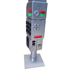 Può includere: Una scultura di robot argentato dal design retrò, con dettagli rossi e verdi. Il robot ha un corpo rettangolare con pulsanti e un piccolo schermo. Poggia su una base bianca. Il design ricorda la fantascienza vintage.