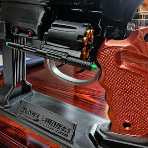 Peut inclure: Une réplique imprimée en 3D d'un pistolet futuriste noir avec une poignée texturée couleur cuivre, un barillet rotatif contenant des cartouches dorées et un support noir portant le texte en relief BLADE RUNNER.
