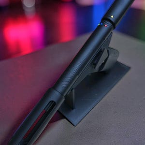 Peut inclure: Un appareil cylindrique noir mat avec une finition texturée, monté sur un support noir. L'appareil présente une série de petits points colorés en vert, bleu et rouge près du sommet, et une longue fente verticale révélant un composant intérieur rouge.