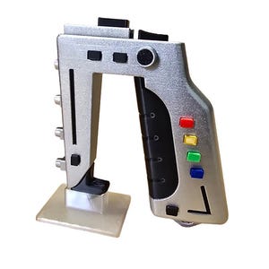 May include: A silver prop gun with a black handle and a square base. The gun has several black buttons and coloured buttons, including red, yellow, and blue. The design is futuristic and detailed.