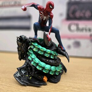 May include: A Spider-Man figurine in a dynamic pose, atop a detailed, multi-layered base with green and black accents. The figure is predominantly red and blue, with a white spider emblem on the chest. The base has a textured, armored appearance.