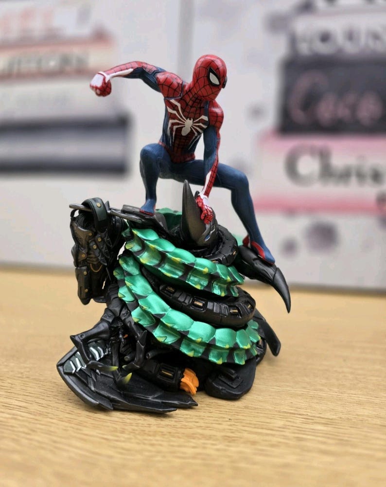 May include: A Spider-Man figurine in a dynamic pose, atop a detailed, multi-layered base with green and black accents. The figure is predominantly red and blue, with a white spider emblem on the chest. The base has a textured, armored appearance.