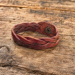May include: Braided leather bracelet in a rich, reddish-brown hue. The bracelet features a woven design and a metal snap closure. The rustic accessory is displayed on a wooden surface, showcasing its texture and craftsmanship.