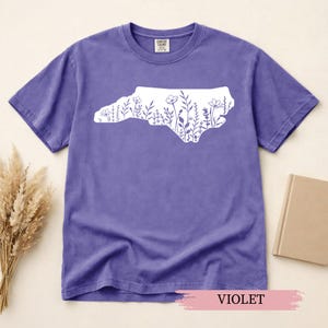 May include: Violet-colored t-shirt featuring a white outline of North Carolina filled with floral designs. The shirt has a crew neck and short sleeves. The text "VIOLET" is displayed at the bottom of the image.