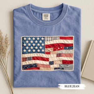 May include: A blue jean colored t-shirt with a sewn-on American flag design. The flag features white stars on a blue background and red and white stripes made of various patterned fabrics. The text "BLUE JEAN" is at the bottom of the image.
