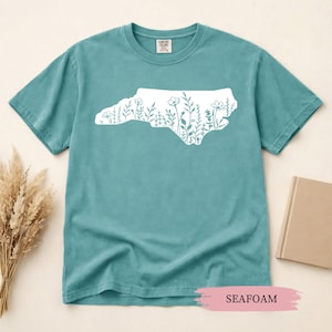 May include: Seafoam green t-shirt featuring a white outline of North Carolina filled with floral designs. The shirt is made of a soft material and has a classic crew neck. The word "SEAFOAM" is printed on a pink background.
