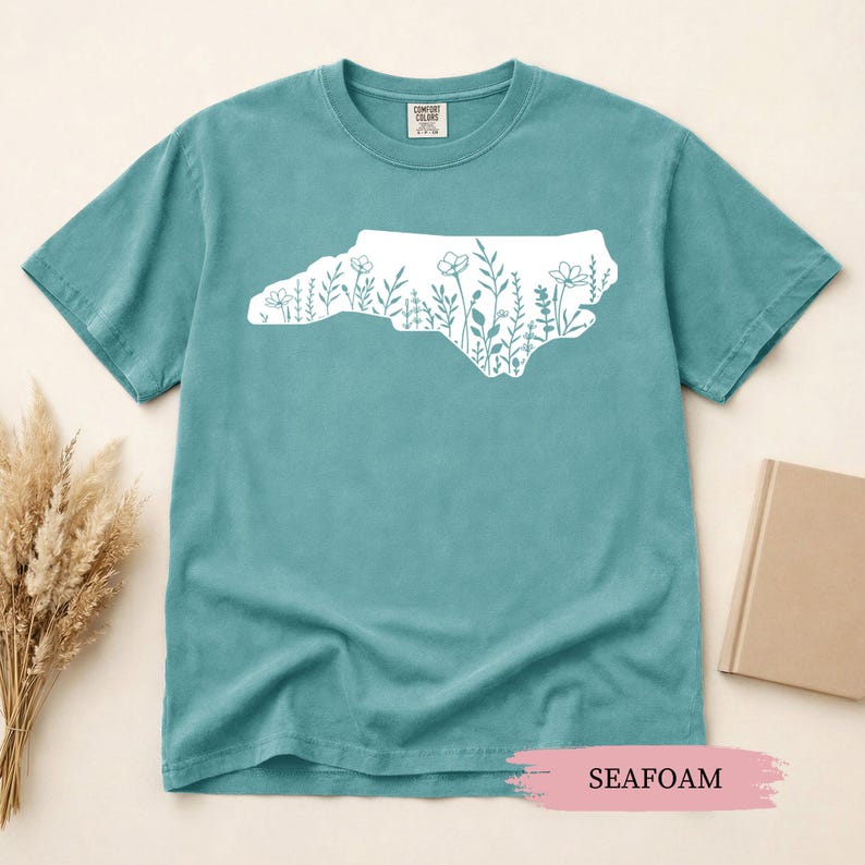 May include: Seafoam green t-shirt featuring a white outline of North Carolina filled with floral designs. The shirt is made of a soft material and has a classic crew neck. The word "SEAFOAM" is printed on a pink background.