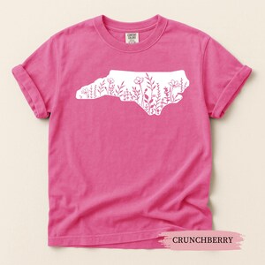 May include: A bright pink t-shirt with rolled-up sleeves. The shirt features a white outline of North Carolina filled with floral designs. The text "CRUNCHBERRY" is at the bottom of the image.
