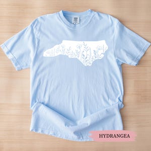 May include: A light blue t-shirt featuring a white outline of North Carolina filled with floral designs. The word "HYDRANGEA" is printed on a pink banner at the bottom of the image. The shirt is made of soft material.