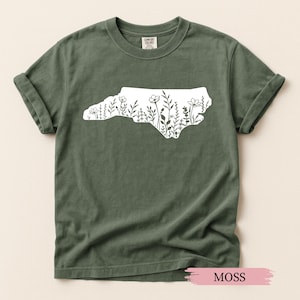 May include: Moss green t-shirt featuring a white outline of North Carolina filled with floral designs. The shirt is made of a soft material and has rolled-up sleeves. The word "MOSS" is printed on a pink label at the bottom.