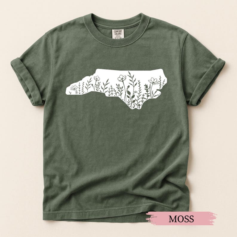 May include: Moss green t-shirt featuring a white outline of North Carolina filled with floral designs. The shirt is made of a soft material and has rolled-up sleeves. The word "MOSS" is printed on a pink label at the bottom.