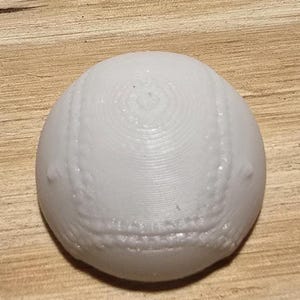 May include: A white, spherical object with a textured surface and stitched detailing, resembling a baseball. The ball is resting on a light brown wooden surface. The stitching is visible around the ball.
