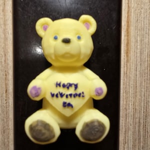 May include: A yellow teddy bear figurine with blue eyes, purple accents, and the text "Happy Veterans' Day" on a heart-shaped chest. The bear is set against a dark rectangular background.