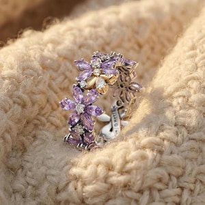 Peut inclure: Une bague en argent ornée d'un motif floral avec des pierres précieuses violettes et transparentes. La bague présente un motif complexe avec un groupe de fleurs. Les pierres précieuses sont serties de manière à capter la lumière, créant un effet scintillant.