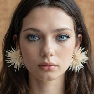 May include: A pair of large, cream-colored, spiky, starburst-shaped earrings worn on the earlobes. The earrings feature numerous thin, pointed, radiating petals that create a textured, spherical appearance, contrasting with the dark hair.