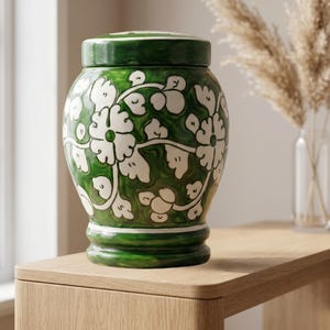 May include: A ceramic jar with a lid, featuring a glossy deep green glaze and a hand-painted white floral vine pattern. The vessel has a rounded body, a defined base, and a matching removable lid, showcasing a smooth, reflective finish throughout.