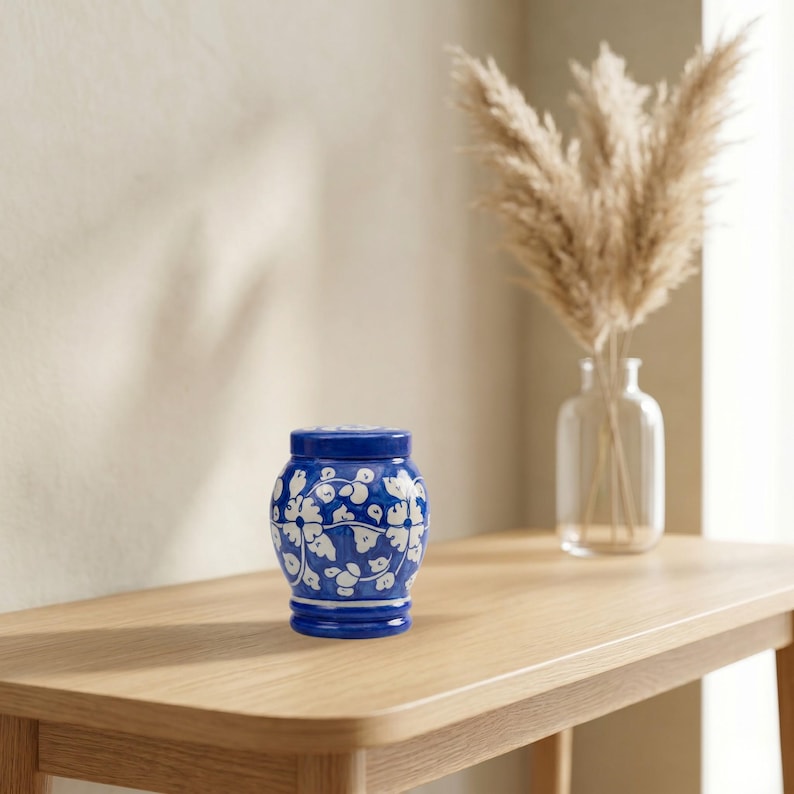 May include: A small, rounded ceramic jar with a lid, featuring a vibrant blue base color and a hand-painted white floral pattern with swirling vines. The jar has a smooth, glossy finish and a distinct, slightly flared base design.