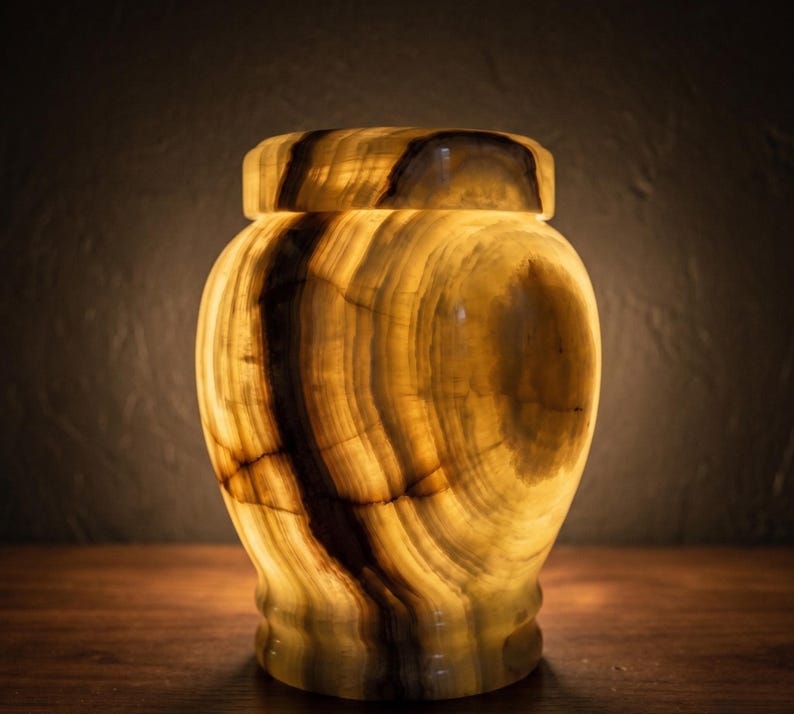 May include: A translucent, bulbous urn crafted from polished stone, featuring warm amber and golden hues with prominent dark brown veining. The vessel has a smooth, rounded body and a matching fitted lid, illuminated from within to highlight the natural banding.