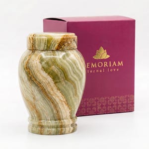 May include: A polished green and cream-colored stone urn with a matching lid, featuring natural swirling patterns. Behind the urn sits a rectangular magenta box with a gold lotus flower logo and the text MEMORIAM eternal love printed on it.