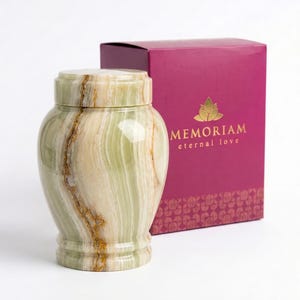 May include: A cylindrical stone urn featuring light green and cream bands with brown veining, topped with a matching lid. Behind the urn sits a rectangular magenta box with the gold text MEMORIAM eternal love and a lotus flower icon.