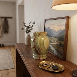 May include: A polished green and brown banded onyx urn with a matching lid, featuring natural stone veining and a smooth, glossy finish. The cylindrical vessel has a slightly flared base and a rounded top, showcasing unique mineral patterns.