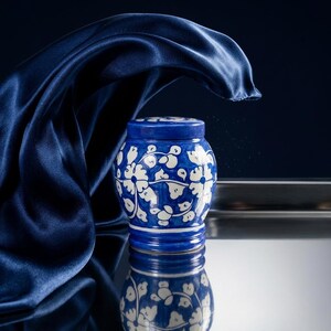 May include: A small, round ceramic jar with a lid, featuring a deep blue glaze decorated with a white floral vine pattern. The jar has a smooth, glossy finish and is positioned against a dark background with a draped navy blue fabric element.