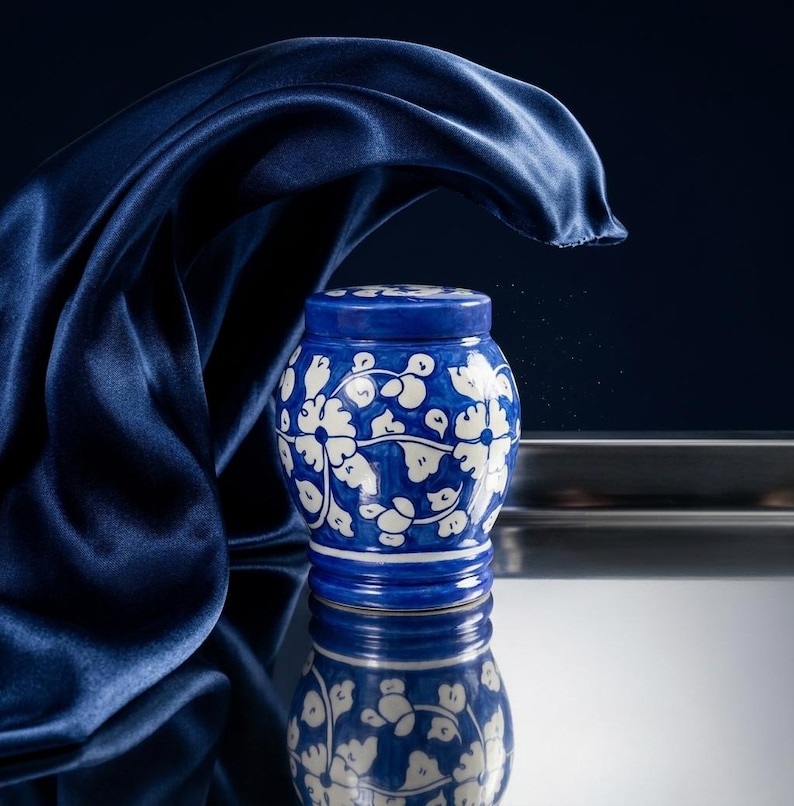 May include: A small, round ceramic jar with a lid, featuring a deep blue glaze decorated with a white floral vine pattern. The jar has a smooth, glossy finish and is positioned against a dark background with a draped navy blue fabric element.