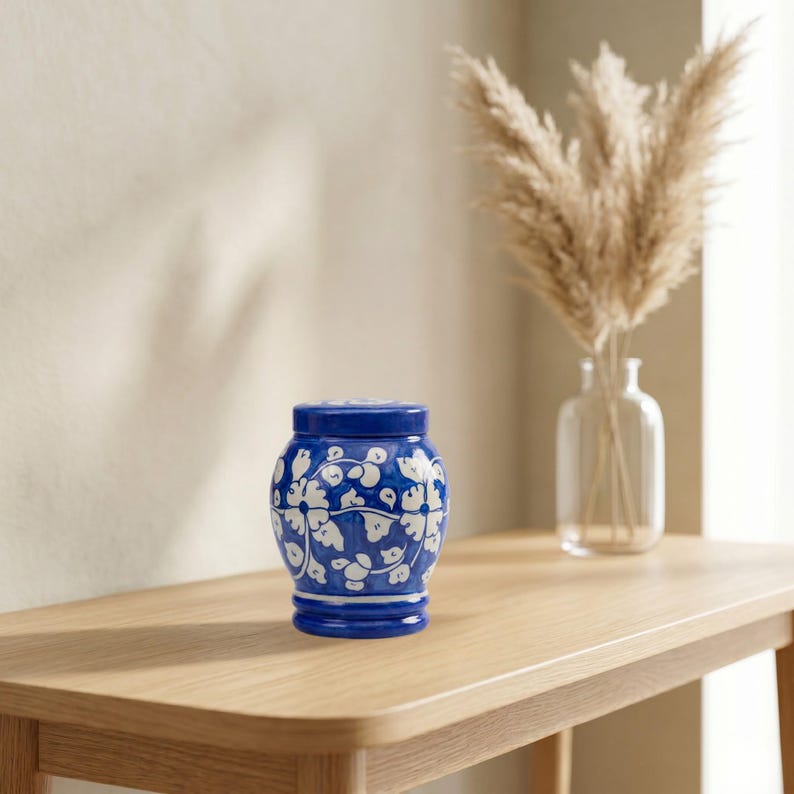 May include: A small, rounded ceramic jar with a lid, featuring a deep blue glaze decorated with hand-painted white floral and vine patterns. The jar has a wide body that tapers toward the base and a matching blue lid with a flat top surface.