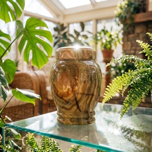 May include: A polished, cylindrical stone urn featuring swirling patterns of brown, tan, and cream colors. The vessel has a rounded body, a slightly flared base, and a matching removable lid with a smooth, reflective finish that catches the light.