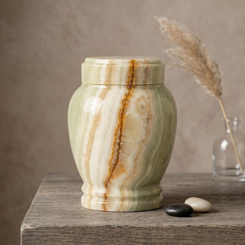 May include: A polished green and cream-colored stone urn with a cylindrical lid and a smooth, banded texture. The vessel features natural mineral veining in shades of amber and white, creating a distinct layered pattern across its rounded body.