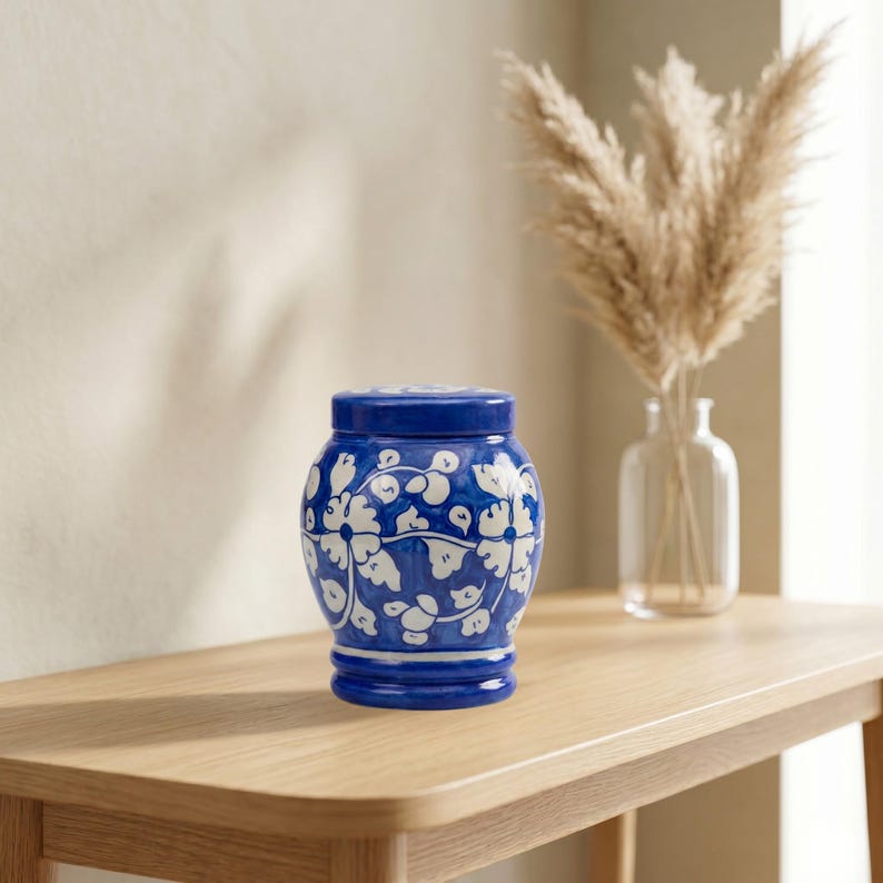 May include: A ceramic jar with a lid, featuring a rounded body and a deep blue base color. The surface is decorated with a hand-painted white floral pattern consisting of stylized blossoms and leafy vines wrapping around the entire exterior.