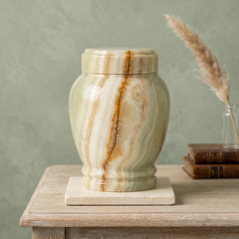 May include: A polished light green and cream-colored stone urn with a matching lid, featuring natural banded patterns and a smooth, glossy finish. The vessel sits on a square, textured beige stone base, showcasing organic mineral veining.