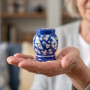 May include: A small, rounded ceramic jar with a lid, featuring a deep blue glaze decorated with a white hand-painted floral and vine pattern. The vessel has a wide body that tapers toward the base and a matching circular lid on top.