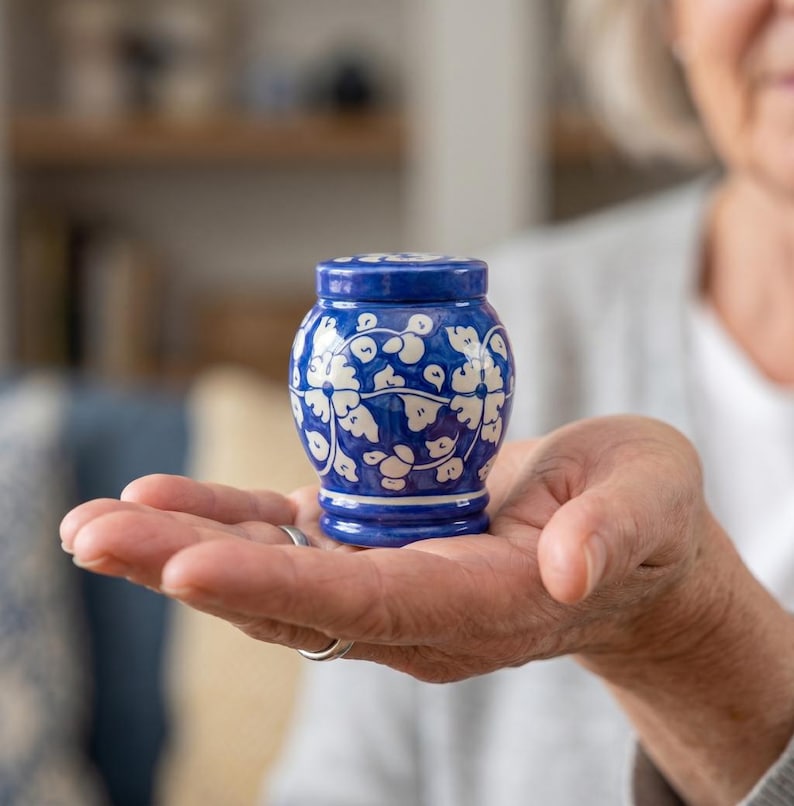 May include: A small, rounded ceramic jar with a lid, featuring a deep blue glaze decorated with a white hand-painted floral and vine pattern. The vessel has a wide body that tapers toward the base and a matching circular lid on top.