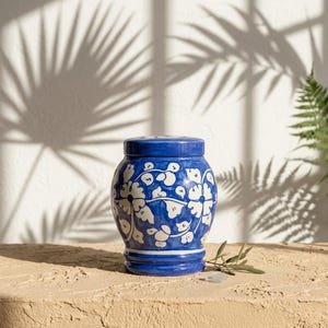 May include: A ceramic jar with a lid, featuring a vibrant blue base color and a hand-painted white floral pattern. The vessel has a rounded body, a defined neck, and a flared base, showcasing a glossy finish with intricate botanical motifs.