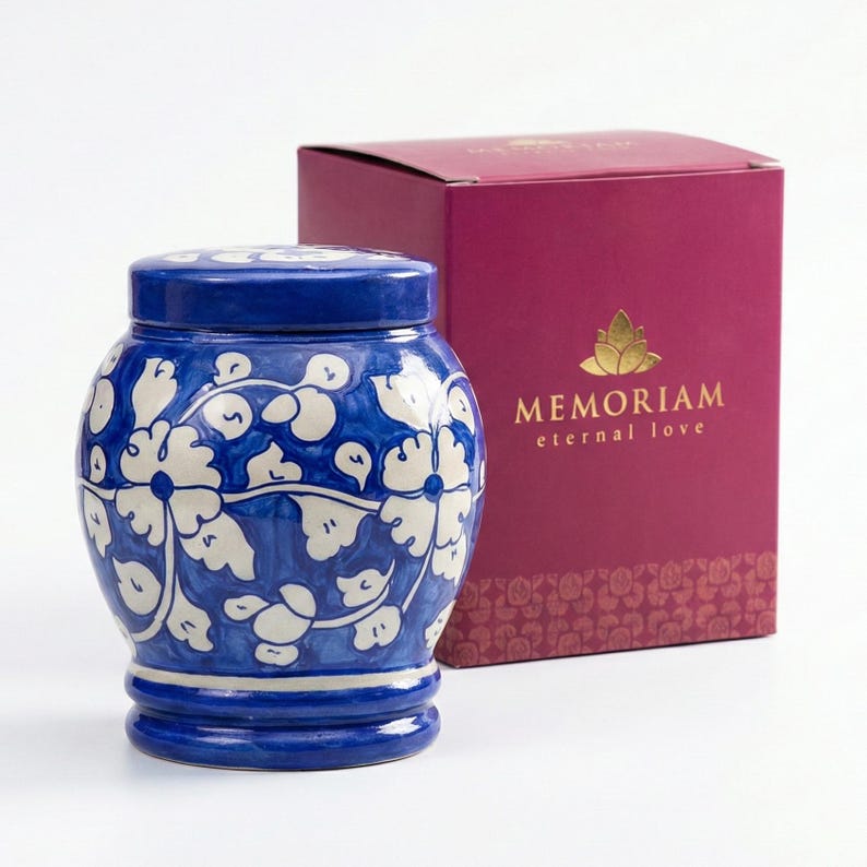 May include: A blue and white ceramic urn with a floral pattern and a matching lid, positioned next to a dark pink rectangular box featuring the gold text MEMORIAM eternal love and a gold lotus flower icon on the front panel of the packaging.