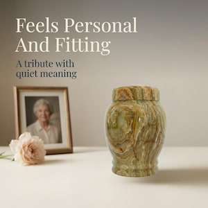 May include: A polished green and brown banded stone urn with a matching lid. The text reads: Feels Personal And Fitting. A tribute with quiet meaning. A framed photograph of a person and a pale pink flower are positioned in the background.
