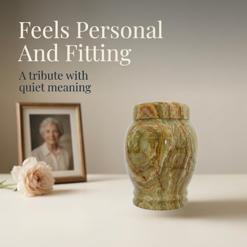 May include: A polished green and brown banded stone urn with a matching lid. The text reads: Feels Personal And Fitting. A tribute with quiet meaning. A framed photograph of a person and a pale pink flower are positioned in the background.