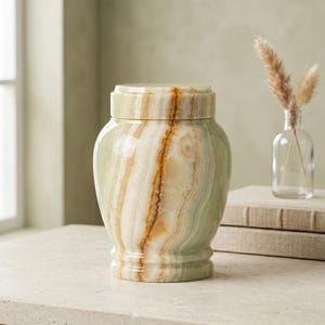 May include: A polished stone urn featuring a smooth, cylindrical body with a matching lid. The surface displays natural, swirling bands of pale green, cream, and warm amber tones, creating a unique, organic pattern across the entire exterior.