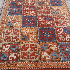 Könnte beinhalten: Ein rechteckiger, handgewebter Teppich mit geometrischem Muster. Der Teppich weist ein sich wiederholendes Design aus Quadraten in Rot-, Blau-, Orange- und Beigetönen auf. Der Rand ist eine Mischung aus Orange und Beige mit einem Fransenrand. Der Teppich ist wahrscheinlich aus Wolle.