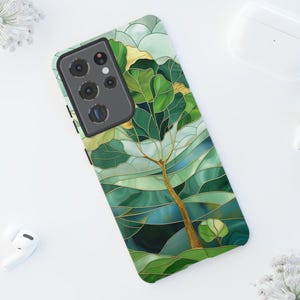 Può includere: Una custodia per smartphone con un design a vetrata raffigurante un albero con foglie verdi e un tronco dorato. La custodia ha un modulo fotocamera nero, circondata da accessori e fiori bianchi.