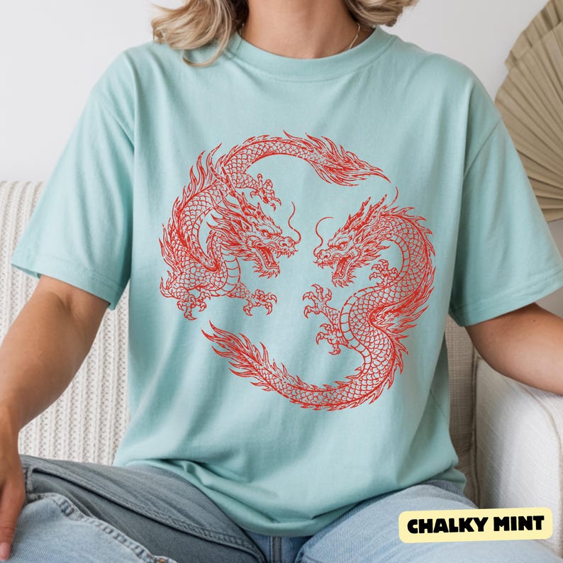 May include: A light mint green short-sleeve t-shirt featuring a large, detailed red graphic print of two dragons intertwined in a circular formation on the chest. The fabric has a smooth texture and the design is centred on the front of the garment.