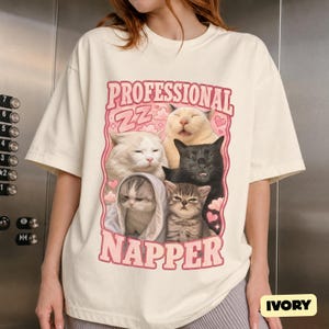 Könnte beinhalten: Ein elfenbeinfarbenes Kurzarm-T-Shirt mit einem Grafikdruck von fünf Katzen sowie dem Text "PROFESSIONAL NAPPER" und "ZZ" in Pink. Die Katzen sind in verschiedenen Schlafpositionen dargestellt, umgeben von rosa Wolken und Herzen.